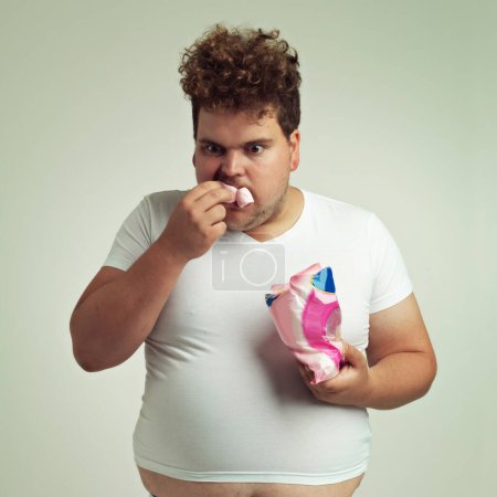 Photo for Food, hungry and marshmallow with plus size man in studio on gray background for unhealthy eating. Hunger, appetite for sweets and ravenous young person with snack bag or packet for greed or gluttony. - Royalty Free Image