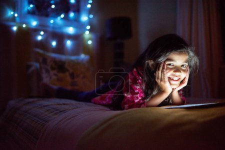 Téléchargez les photos : Nuit, sourire et tablette avec portrait de fille dans la chambre à coucher de la maison pour jouer ou regarder un film. Lit, internet et technologie avec un jeune enfant heureux dans l'appartement pour la navigation en soirée ou de divertissement. - en image libre de droit