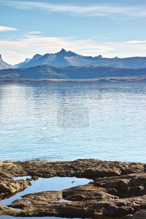 Téléchargez les photos : Lac, paysage et montagnes dans la nature avec des roches en Norvège sur le rivage avec écologie pour l'environnement. Eau, mer ou océan avec colline, pierre et plein air avec nuages à l'horizon pour la durabilité. - en image libre de droit