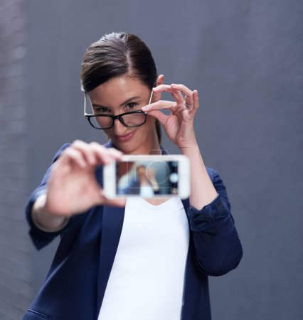 Photo for Businesswoman, selfie and cellphone by dark background for online company, social media and pride. Confident female person, technology and glasses with emoji face for internet and profile picture. - Royalty Free Image