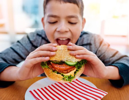 Téléchargez les photos : Garçon, maison et manger un hamburger pour le déjeuner, la nutrition et la restauration rapide délicieux pour le développement de l'enfance. Affamé, personne masculine avec un délicieux repas de glucides, des condiments sains et morsure pour le bien-être et la croissance. - en image libre de droit