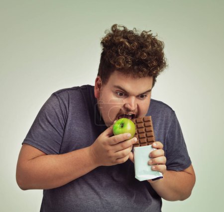 Photo for Person, apple and chocolate in studio with choice of junk food, sweet or diet for wellness. Plus size, man eating and decision, fruit or candy for nutrition, lose weight and healthy lifestyle. - Royalty Free Image