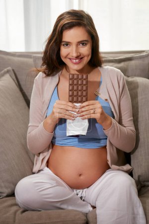 Foto de Sonrisa, chocolate y retrato de la mujer embarazada en el sofá para relajarse con merienda en la sala de estar en el apartamento. Feliz, maternidad y persona femenina anhelando postre dulce en el sofá en el salón del hogar - Imagen libre de derechos