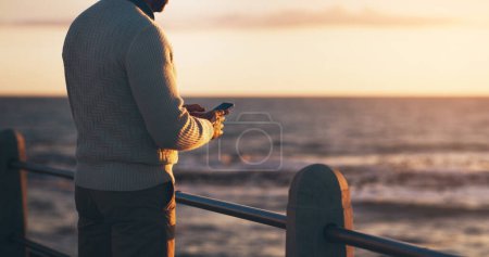 Homme, téléphone et textos sur la promenade par l'océan avec coucher de soleil ciel en vacances avec contact sur le web. Personne, smartphone et extérieur par la mer, vagues et retour en vacances à l'horizon avec application pour les médias sociaux.