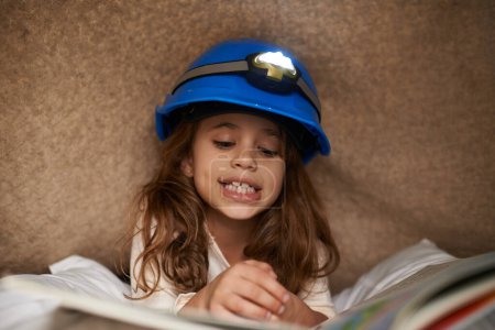 Photo for Girl, child and book with headlamp for reading, above and learning with smile for education in family home. Kid, story and happy with light for info, language and literacy skills on floor in house. - Royalty Free Image