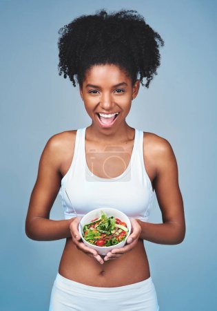 Foto de Sonríe, retrata o mujer negra con ensalada en estudio para una comida saludable, dieta nutricional o beneficios de digestión. Cuerpo, verduras y feliz modelo de fitness femenino comiendo alimentos aislados sobre fondo azul. - Imagen libre de derechos