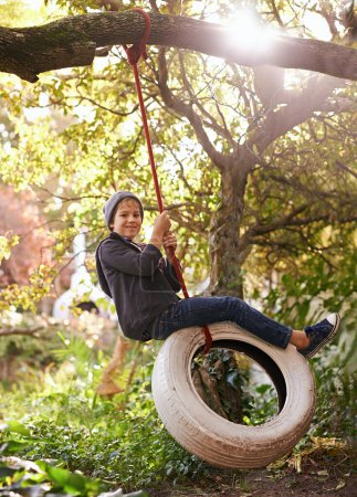 Boy, child and tyre swing with portrait in garden with smile, playing and countryside vacation in summer. Kid, face and diy adventure playground in backyard of home with sunlight and trees in nature.