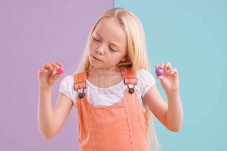 Photo for Studio, girl and decision with candy, dessert and choice for snack and childhood. Child, gumball and yummy selection for tasty, eating and choosing on vibrant split pastel pink and blue background. - Royalty Free Image
