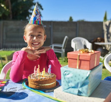 Téléchargez les photos : Fête, portrait et enfant avec gâteau d'anniversaire, présent et bonheur à l'événement en plein air dans le jardin. Excité, enfant et célébration à table dans le parc en vacances, en vacances et merci pour la nourriture et le cadeau. - en image libre de droit