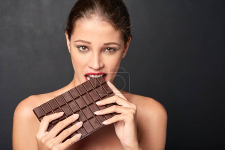 Chocolate, portrait and woman in studio eating for unhealthy diet, craving sugar or cheat day on dark background. Bite, face and female model for snack temptation, sweet addiction or cacao candy bar.