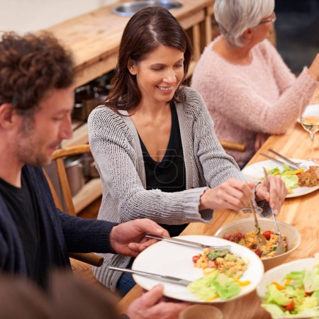 Photo for Family, people and lunch in home dining room with food, happy and relax with bonding, connection or event. Man, women and smile for meal, diet and nutrition on holiday, plate and help giving salad. - Royalty Free Image