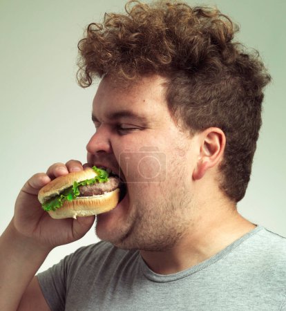 Foto de Obesidad, aumento de peso y comida rápida para el hombre en el estudio con hamburguesa para comer poco saludable, además de tamaño y codicia. Persona con sobrepeso con hamburguesa en la boca y barbilla doble para deliciosa, humor y comedia. - Imagen libre de derechos
