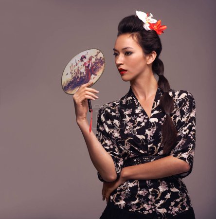 Foto de Mujer japonesa, moda y abanico tradicional con confianza en estudio sobre fondo marrón para cultura, estilo y elegante. Mujer, camisa y flor en la cabeza con ropa de diseñador o atuendo. - Imagen libre de derechos