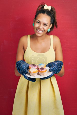 Photo for Happy woman, portrait and baker with cupcakes for dessert, food or sweet treat on a red studio background. Young female person with smile or mittens for hot baking or hospitality in yellow dress. - Royalty Free Image