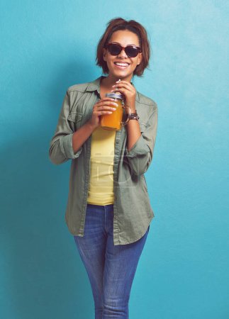 Foto de Mujer feliz, retrato y gafas de sol con zumo de naranja de moda para vitamina C o dieta en un fondo de estudio azul. Mujer, morena o modelo con sonrisa para bebida cítrica o bebida fresca. - Imagen libre de derechos