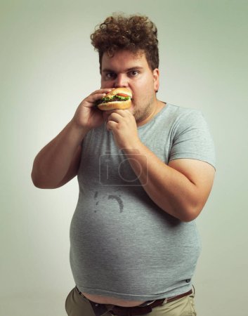 Burger, plus la taille et le portrait pour l'homme en studio avec un régime alimentaire pour une alimentation malsaine, affamée et avide. Jeune avec repas dans la bouche et l'estomac rond pour mangeur désordonné, calories et à emporter à l'intérieur.