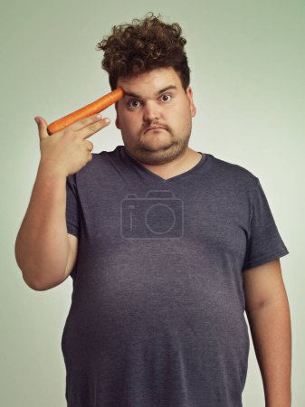 Photo for Portrait, carrot and hand gun with plus size man in studio on gray background for diet, health or nutrition. Depression, sad and vegetable to head of young person in conflict with vegetarian food. - Royalty Free Image