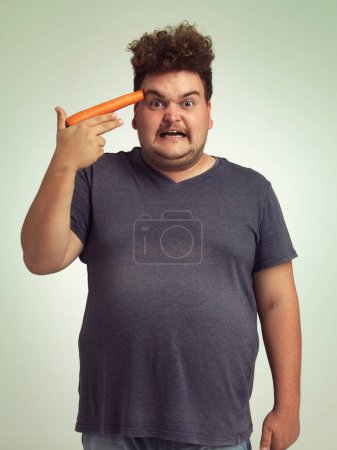 Photo for Portrait, vegetable and hand gun with plus size man in studio on gray background for diet, health or nutrition. Anger, depression and carrot to head of young person in conflict with vegetarian food. - Royalty Free Image