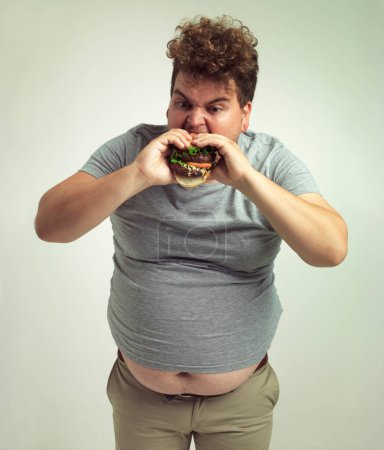 Foto de Obesidad, aumento de peso y hamburguesa para el hombre en el estudio con comida para llevar para comer poco saludable, además de tamaño y codicia. Persona con sobrepeso con comida en boca y estómago gordo para el hambre, la comedia y la mordedura en interiores. - Imagen libre de derechos