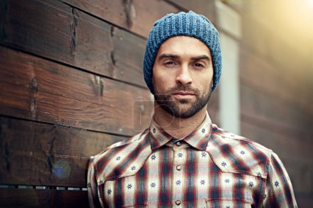 Photo for Portrait, confident and man on wood wall background outdoor in casual clothes, outfit or apparel. Face, serious and stylish person in beanie, shirt and trendy fashion for profile picture in Spain. - Royalty Free Image