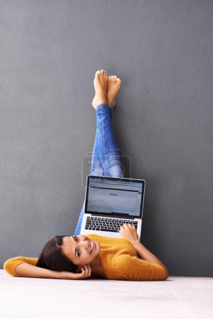 Die Vorteile der Technologie. Eine junge Frau liegt mit ihrem Laptop vor grauem Hintergrund auf dem Boden