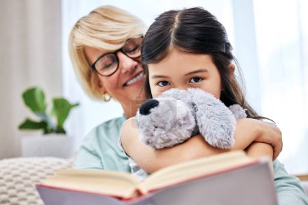 Photo for Smile, book and senior woman with child reading for knowledge with teddy bear at home together. Happy, bonding and elderly grandmother relaxing and enjoying story with girl kid holding toy at house - Royalty Free Image