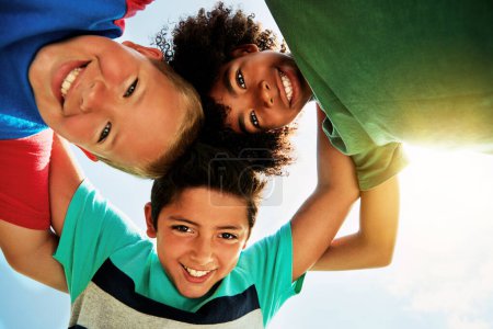 Photo for Happy, blue sky and low angle portrait of children for friendship, playing and fun together. Diversity, youth and boys huddle in circle for childhood, game and hug for school, recess and playground. - Royalty Free Image