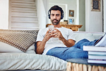 Photo for Man, phone and video with headphones on sofa for sound, audio and streaming service in living room with relax. Person, smartphone and mobile app for listening to music, radio and subscription in home. - Royalty Free Image