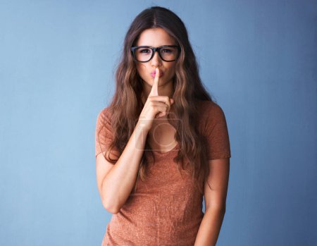 Photo for Girl, glasses and finger for secret on studio backdrop, mock up and hush sale with optometry customer for promotion. News, privacy and gossip, mute and hand gesture on blue background for deal. - Royalty Free Image