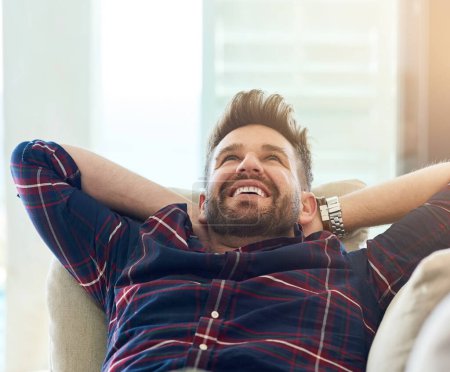 Happy, man and stretching on sofa to relax, rest and energise at home on weekend or vacation. Male person, smile and hands behind head on couch for comfort and wellness on day off or holiday.