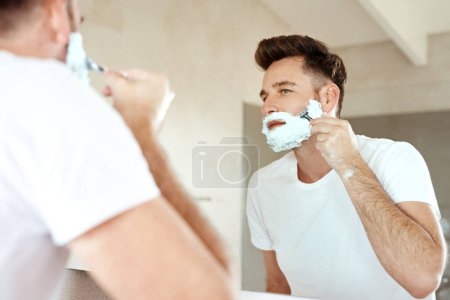 Photo for Man, mirror and shaving face with razor in bathroom for grooming, skincare or morning routine. Reflection, foam and person cutting beard for cleaning, health and hair removal for hygiene in home. - Royalty Free Image