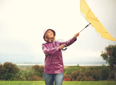 Foto de Upset, woman and umbrella with raincoat for bad weather and rainstorm at park in nature. Female person, rain and fighting with wind for cover or outdoor protection in winter season at Glasgow. - Imagen libre de derechos