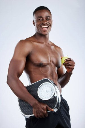 Foto de Hombre africano, fitness y sonrisa con manzana y báscula para adelgazar y dieta equilibrada en estudio. Feliz, hombre o culturista con metas de frutas, nutrición y cuerpo para entrenar y hacer ejercicio. - Imagen libre de derechos