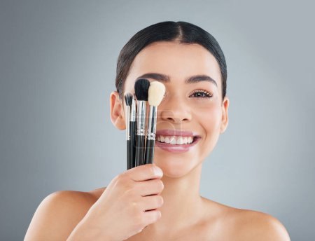 Photo for Young woman, studio and beauty with brushes for facial transformation, skincare or cosmetic. Female person, gray background and smile with tool on eye for wellness, glow and shine in portrait. - Royalty Free Image