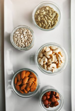 Bowls, nuts and seeds on plate, above and hazelnut for nutrition, health and table with diet for wellness. Cashew, almond and pumpkin in glass container for meal prep with food in morning at house.