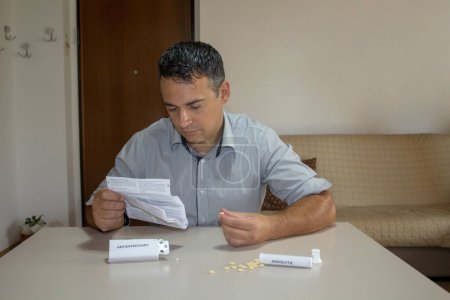 Photo for Man sitting while reading the leaflet of medicines with anxiolytic and antidepressant pills scattered on the table. Problem and consequences of psychotropic drugs. - Royalty Free Image
