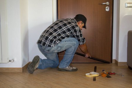 Téléchargez les photos : Un homme à tout faire installant un joint anti-poussière sur la porte de la maison. Meilleure isolation thermique et nettoyage du sol. - en image libre de droit