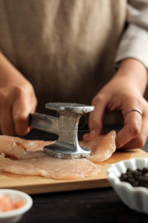 Téléchargez les photos : Femme cuisiner dans la cuisine, battre la viande de poulet en utilisant attendrisseur de viande. Processus de cuisson - en image libre de droit