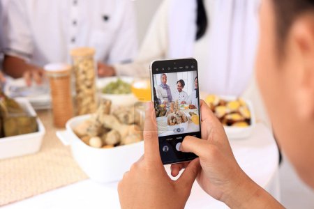 Männliche Hand fotografiert asiatische Familie und Freund, die Eid Mubarak mit Essen auf dem Tisch feiern 