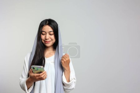Mujer indonesia con velo feliz usando el teléfono móvil, sonrisa emocionada. Eid Festive Happy Concept. Copiar espacio para texto 