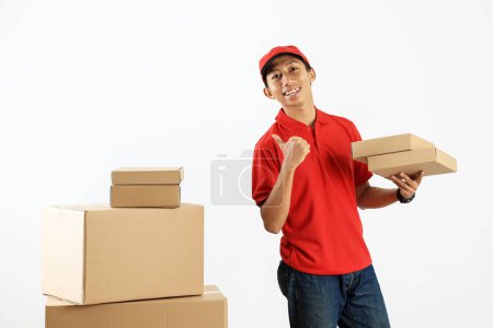 Smiling Asian Courier Delivery Man in Red Uniform Holding Cardboard in Warehouse. Thumb Up Pose 