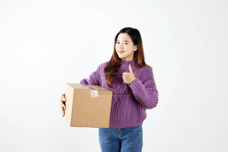 Asiatische junge Frau hält Paketkasten mit Daumen hoch Geste, Online-Shopping-Konzept. Isoliert auf Weiß 