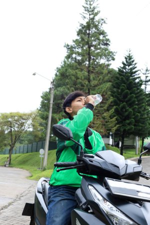 Asiático joven conductor de moto con conductor en línea aplicación chaqueta beber una botella de agua mineral