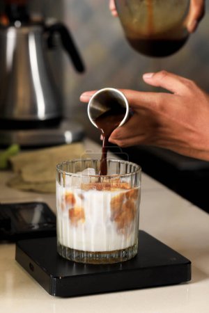 Männliche Hand Barista gießt schwarzen Kaffee Espressoschokolade mit Messbecher ins Glas mit frischer Milch und Eis