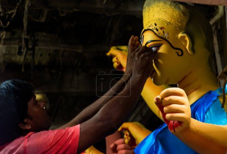 Foto de Kolkata, India - 25 de septiembre de 2022: Un artesano de mediana edad ocupado dentro de su taller en Kumortuli. Está profundamente concentrado en pintar el ojo de una hermosa diosa ídolo de arcilla Durga. Enfoque selectivo. - Imagen libre de derechos