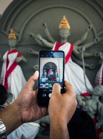 Foto de Un hombre haciendo clic en la foto de la Diosa Durga en el móvil en la Feria del Libro de Calcuta. Enfoque selectivo en el móvil con otro borroso. - Imagen libre de derechos