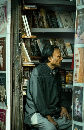 Foto de Kolkata, India - 12 de febrero de 2023: Un viejo librero sentado en medio de estantes de libros, mirando pensativamente a un lado. Enfoque selectivo. - Imagen libre de derechos