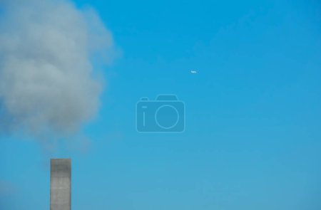 Smoke from a chimney and an airplane against a background of blue sky on a sunny day.