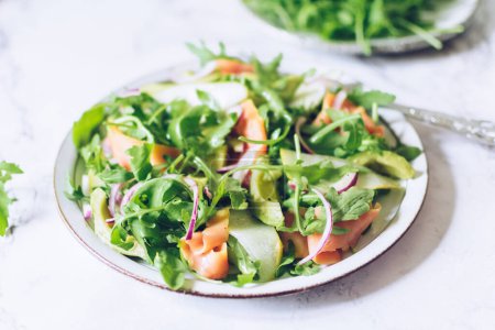 Gesunder Salat mit Rucola, Räucherlachs, Birne, Avocado und roter Zwiebel auf einem weißen Teller auf weißem Marmorgrund. Leckeres vegetarisches Kochbuch. Diät-Fitness-Menü. Ansicht von oben.