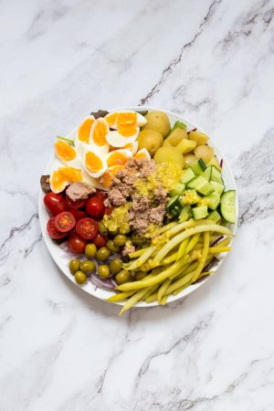 Nicoise Salat - Salat nach französischer Art mit Eiern, Thunfisch, Kirschtomaten, grünen Oliven, Kartoffeln, grünen Bohnen und Gurken auf einem großen Teller auf weißem Marmorgrund. Leckeres Essen Rezept Kochbuch. Ansicht von oben.